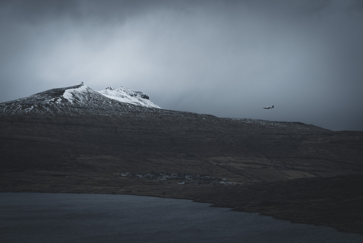Färöer Anflug, Flugzeug über Dorf am Fjord vor schneebedecktem Berg im Wintergrau, Düstere Weite - Landschaftsfotografie