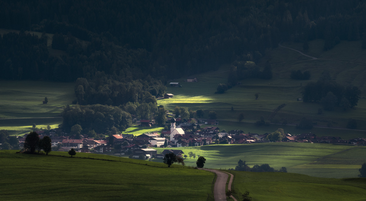 Fischen, Allgäuer Dorf mit Kirchturm im Sonnenlicht zwischen dunklen Berghängen, Licht und Schatten im Tal
