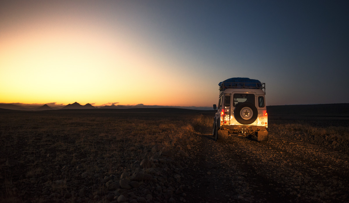 Land Rover Defender auf Schotterpiste, Bergsilhouetten bei Sonnenuntergang, Freiheit im Hochland - Reisefotografie Island