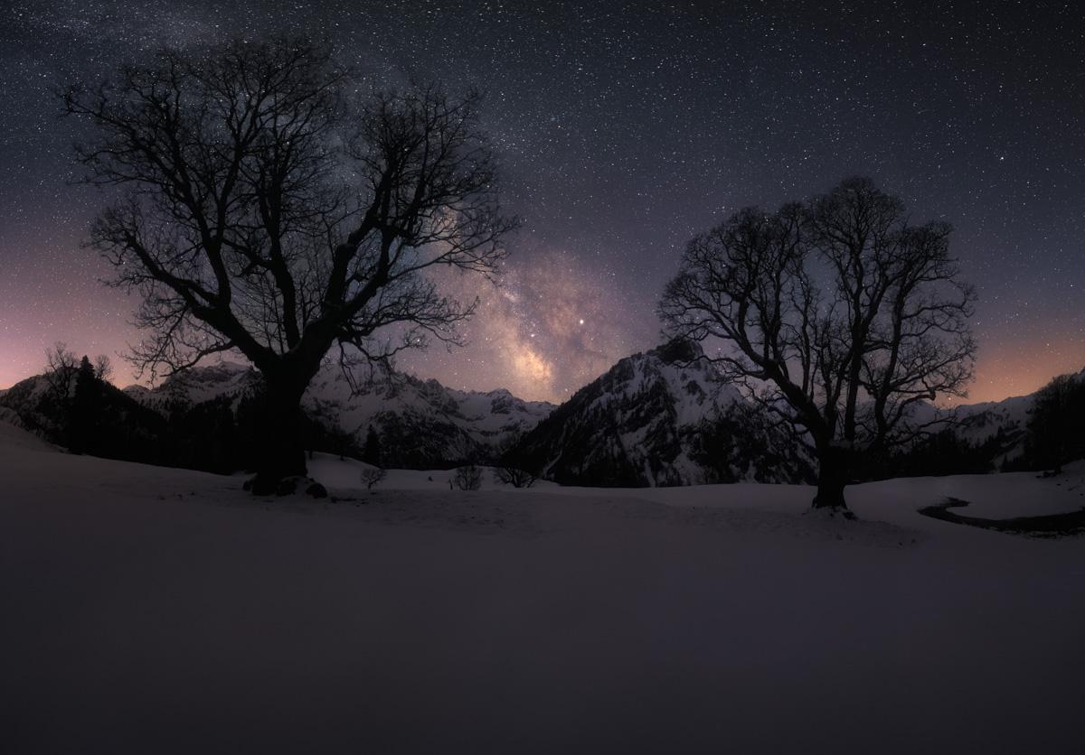 Milchstraße über Hinterstein im Winter, Baumsilhouetten und verschneite Berge unter dem Sternenhimmel, Winterliche