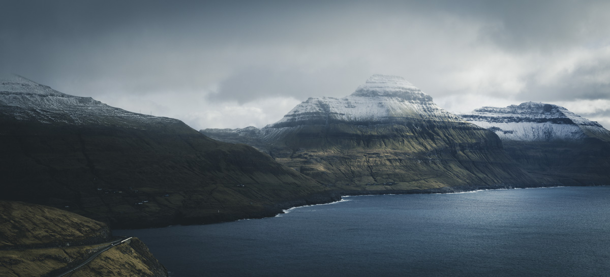 Färöer, Verschneiter Pyramidenberg über düsterem Fjord mit Küstenstraße, Winterliche Berglandschaft - Landschaftsfotografie