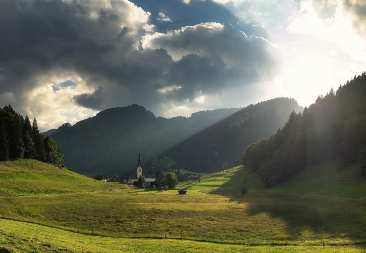 Allgäuer Bergdorf im Sonnenlicht, Kirchturm zwischen grünen Wiesen und dramatischen Wolken, Oberstdorf - Allgäu