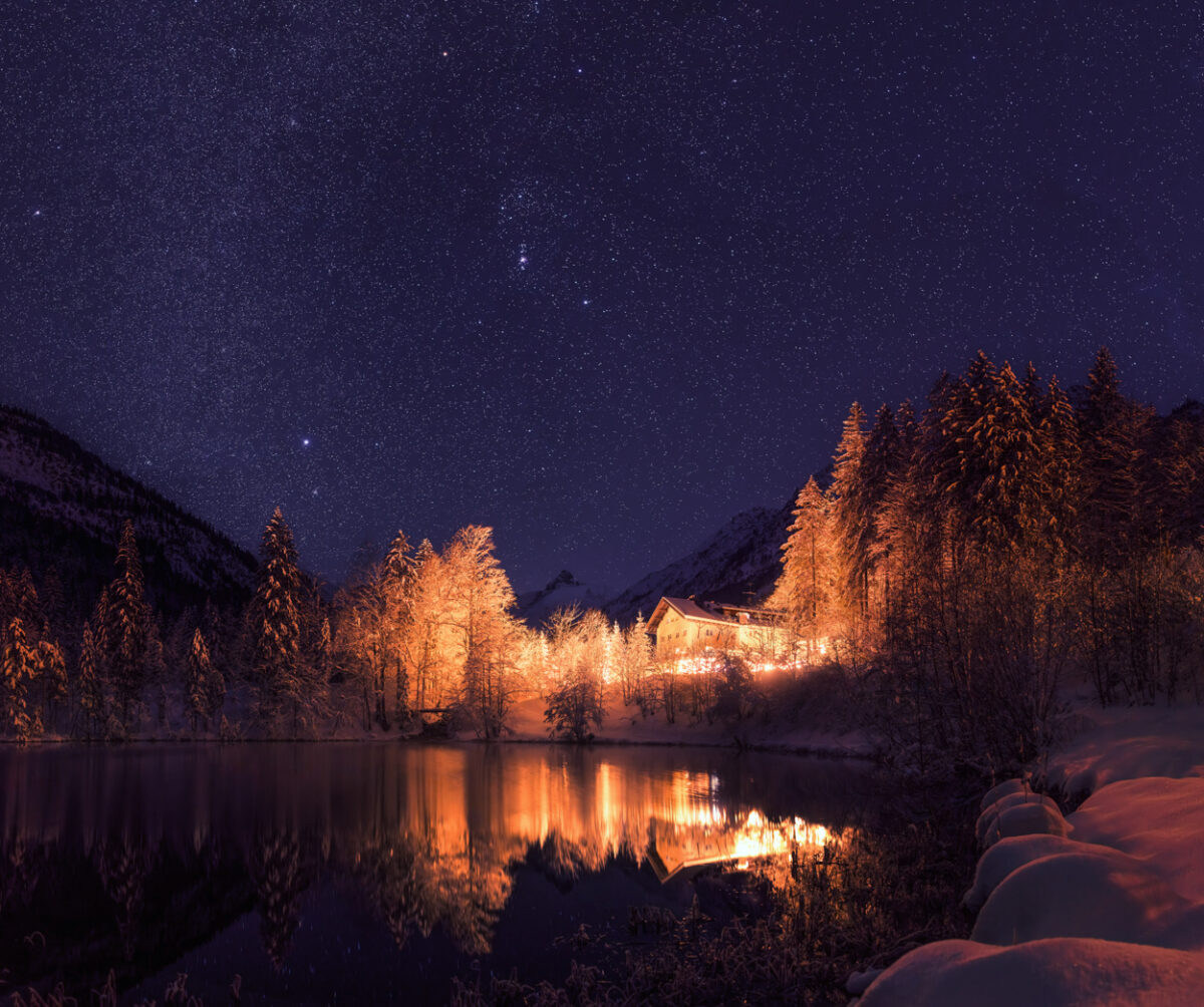 Christlesee Oberstdorf, Winterliche Sternennacht am verschneiten Bergsee, Warme Lichter spiegeln sich im stillen Wasser