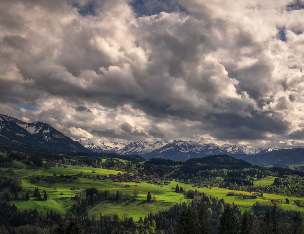 Imberg Frühlingswiesen Panorama, Saftig grüne Almwiesen mit Bergdorf vor dramatischem Wolkenhimmel, Weitblick über