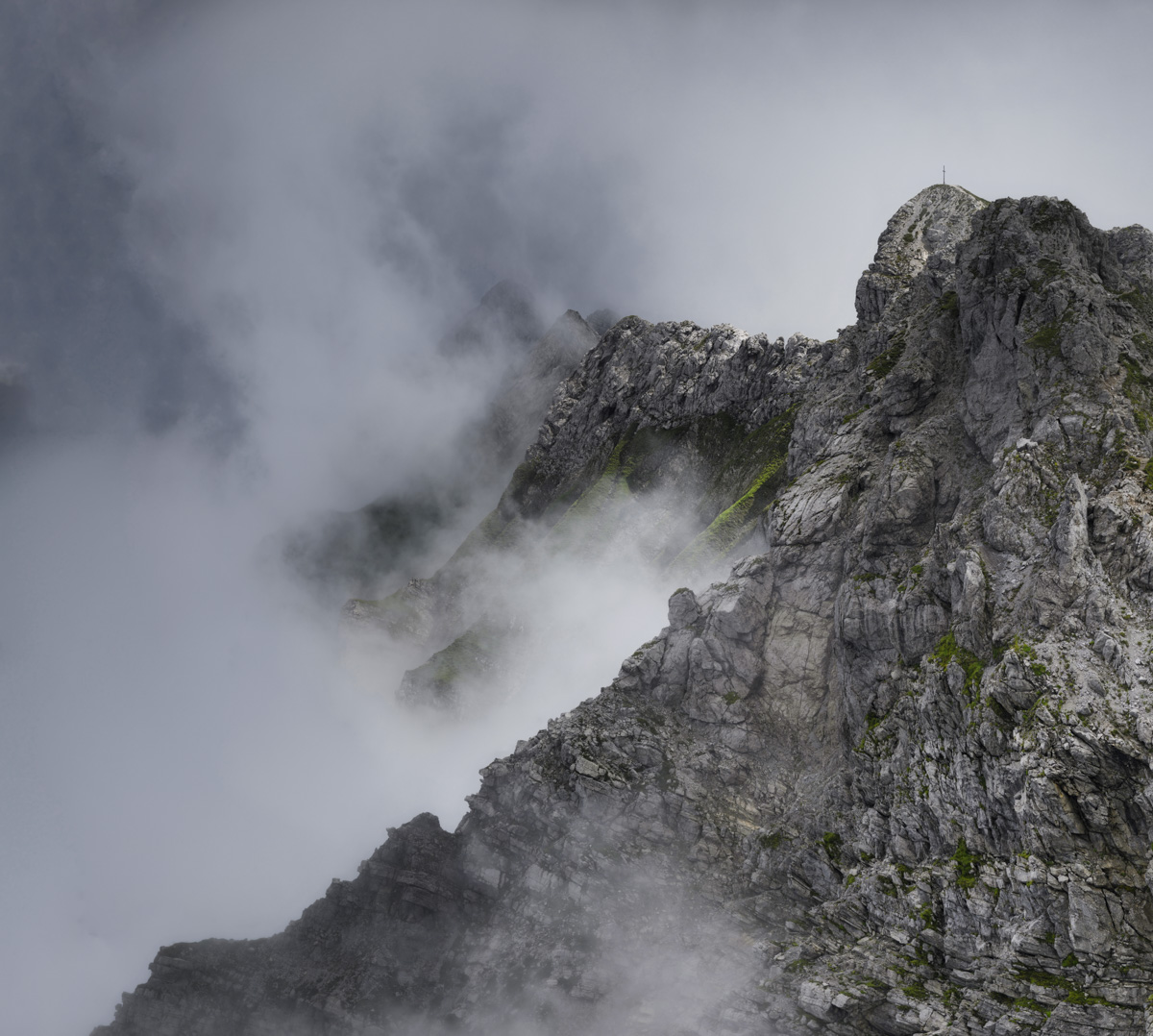 Hindelanger Klettersteig Gipfelkreuz im Nebel, Schroffe Felswand mit Gipfelkreuz zwischen Wolkenfetzen, Dramatische